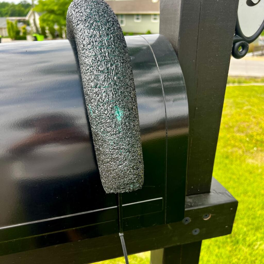 A black foam pool noodle is attached vertically to the curved door of a black mailbox, likely serving as a protective bumper. This DIY patriotic mailbox swag is mounted on a wooden post, with grass and houses visible in the background.