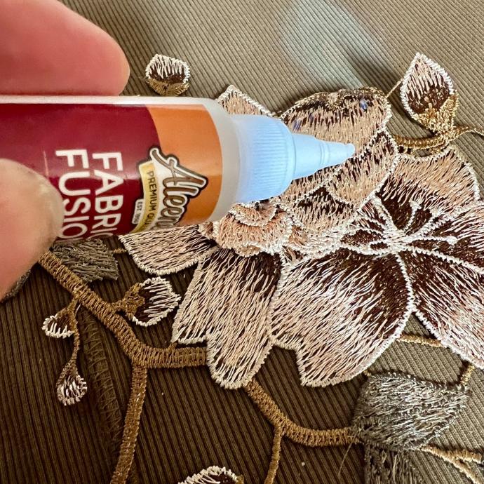 A hand holds a bottle of fabric glue with a blue tip, applying it to a textured floral applique sewn onto a brown fabric background.