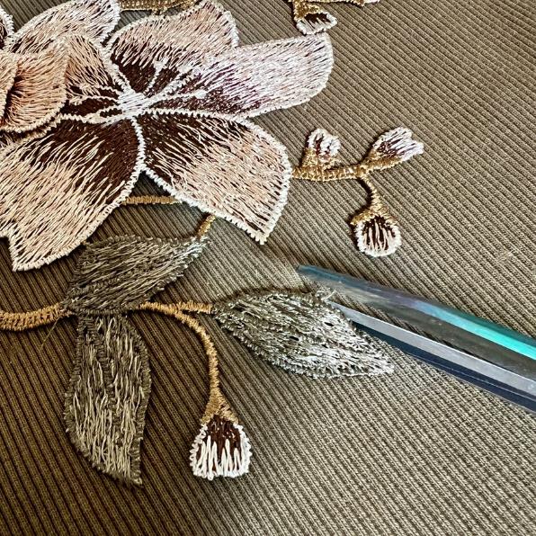 A close-up of scissors cutting around a detailed embroidered flower and leaves on textured fabric, preparing to cut out the embroidery piece.