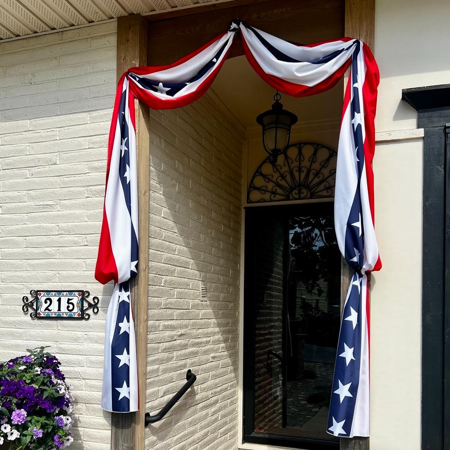 A doorway is decorated with red, white, and blue bunting featuring white stars. A black lantern hangs above the door, and a plaque with the number 215 is mounted on the brick wall next to a pot of purple flowers.