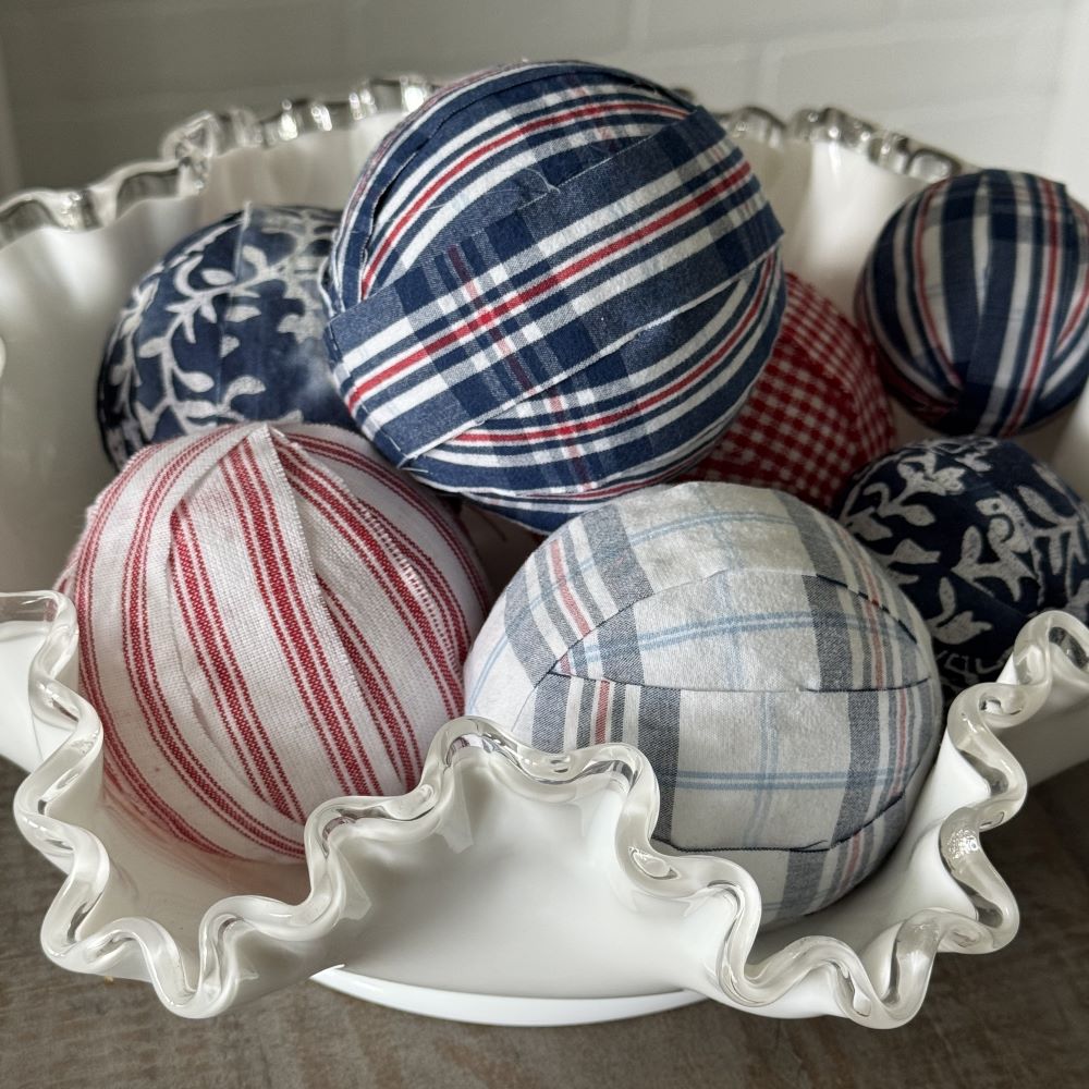 A white ruffled bowl holds decorative fabric-covered balls with red, white, and blue patterns, including stripes, plaid, and floral designs.