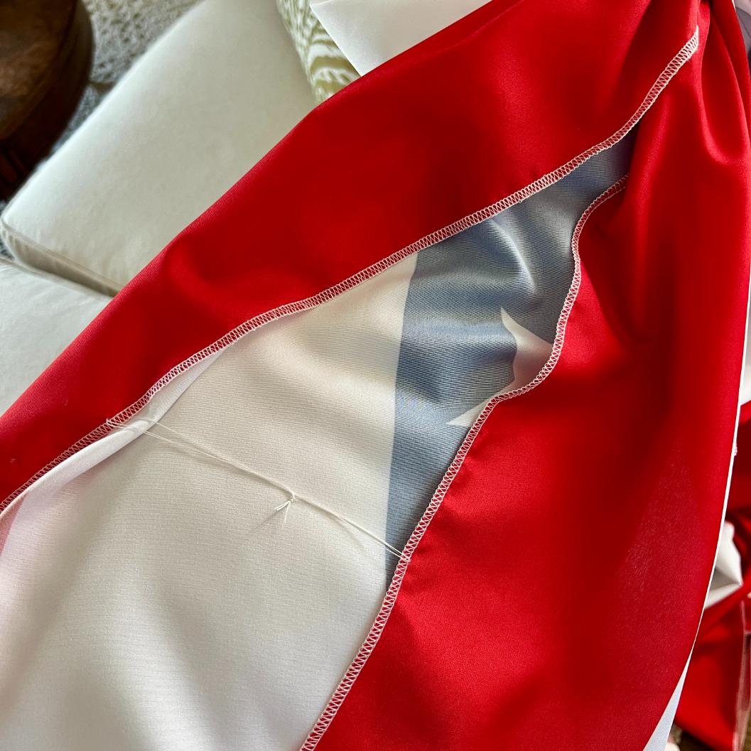 Close-up of fabric with a bright red edge and white center, featuring zigzag stitching along the seam. The material appears draped over a light-colored couch with a cushion in the background.
