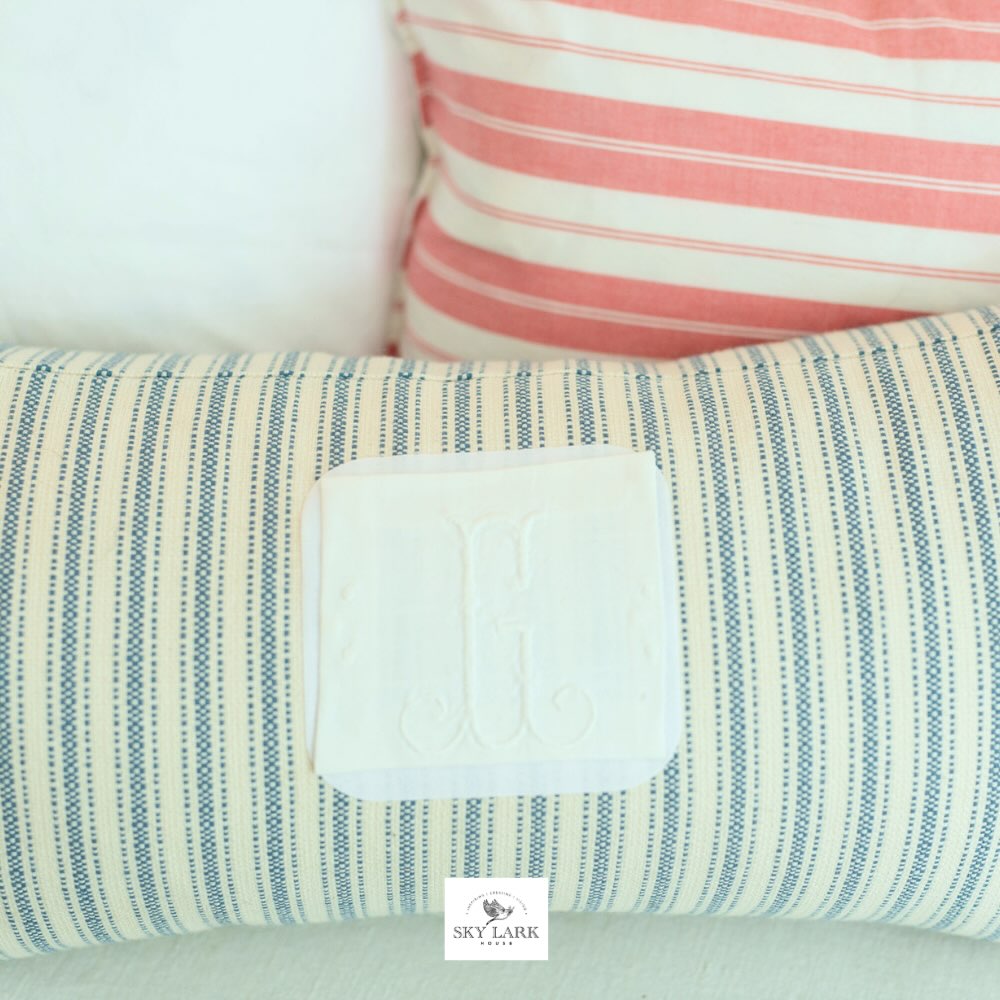A close-up of two pillows: one with pink and white stripes in the background, and one with blue and white stripes and a white monogrammed patch in the foreground. A small "SKY LARK" label is visible at the bottom.