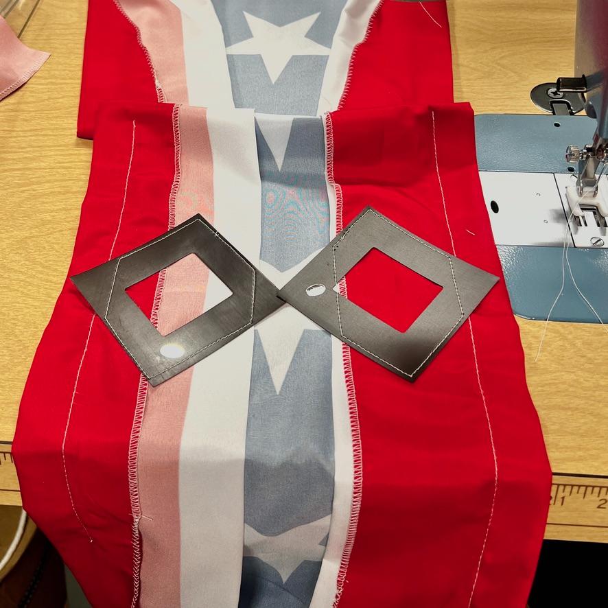 A sewing project features red, white, and blue star-patterned fabric on a table beside a sewing machine, with two black square templates placed on top.