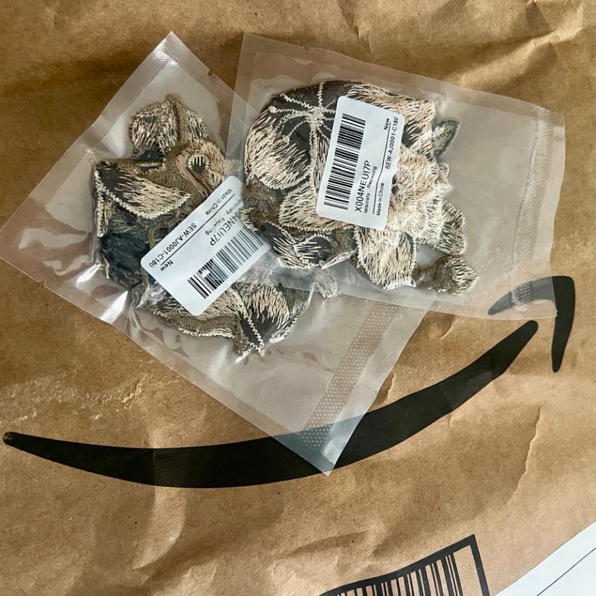 Two embroidered dog patches sealed in plastic bags, each with a barcode sticker, placed on a brown Amazon package with a partial black Amazon logo visible.