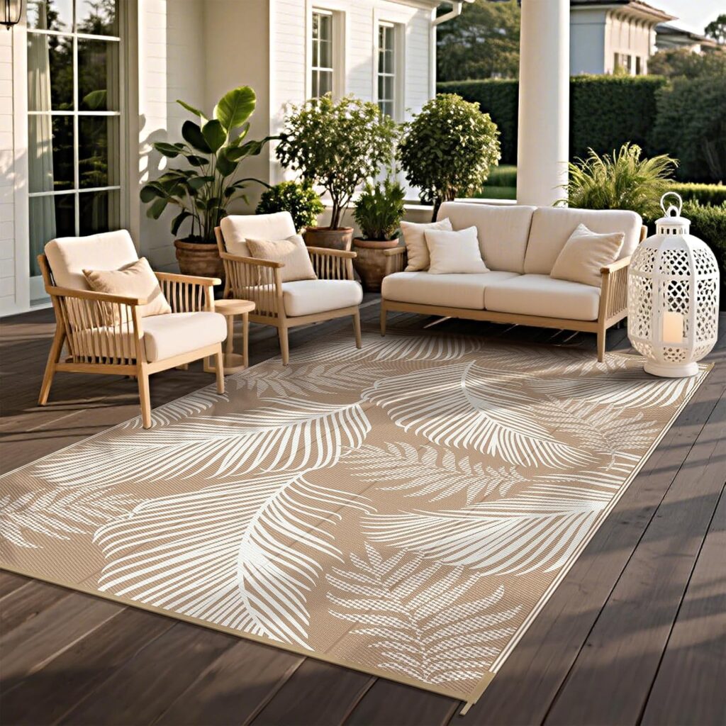 A cozy outdoor patio with cream cushioned chairs, a sofa, potted plants, a large tropical-print rug, and a decorative white lantern, set on a wooden deck beside a white house with columns.