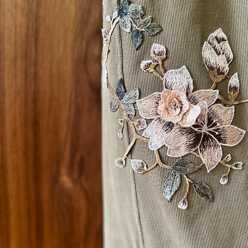 Close-up of beige corduroy fabric with intricate floral embroidery in shades of cream, pink, and gray, set against a blurred wooden background.