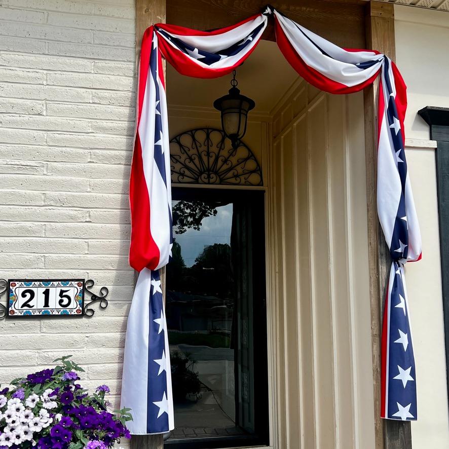 A doorway decorated with red, white, and blue star-spangled bunting, next to a house number sign reading 215 on a white brick wall. Purple flowers are in the foreground.