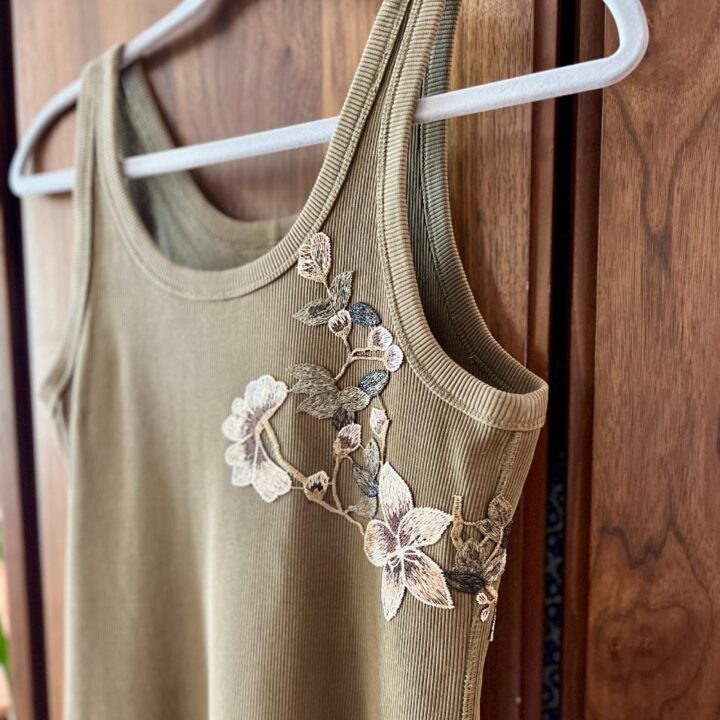 A ribbed olive-green tank top hangs on a white hanger, featuring delicate floral embroidery with cream and brown flowers and leaves on one shoulder and along the neckline. Wooden panels are in the background.