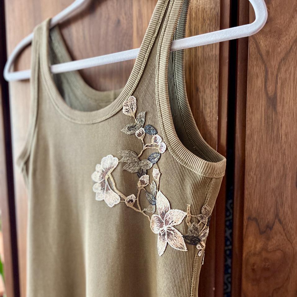 A ribbed olive-green tank top hangs on a white hanger, featuring delicate floral embroidery with cream and brown flowers and leaves on one shoulder and along the neckline. Wooden panels are in the background.