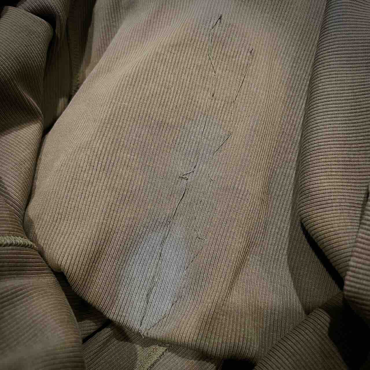 A close-up of a brownish ribbed fabric with noticeable patches and visible stitched repairs, suggesting wear and mending. The fabric appears to be from a piece of clothing, such as a sweater or jacket.