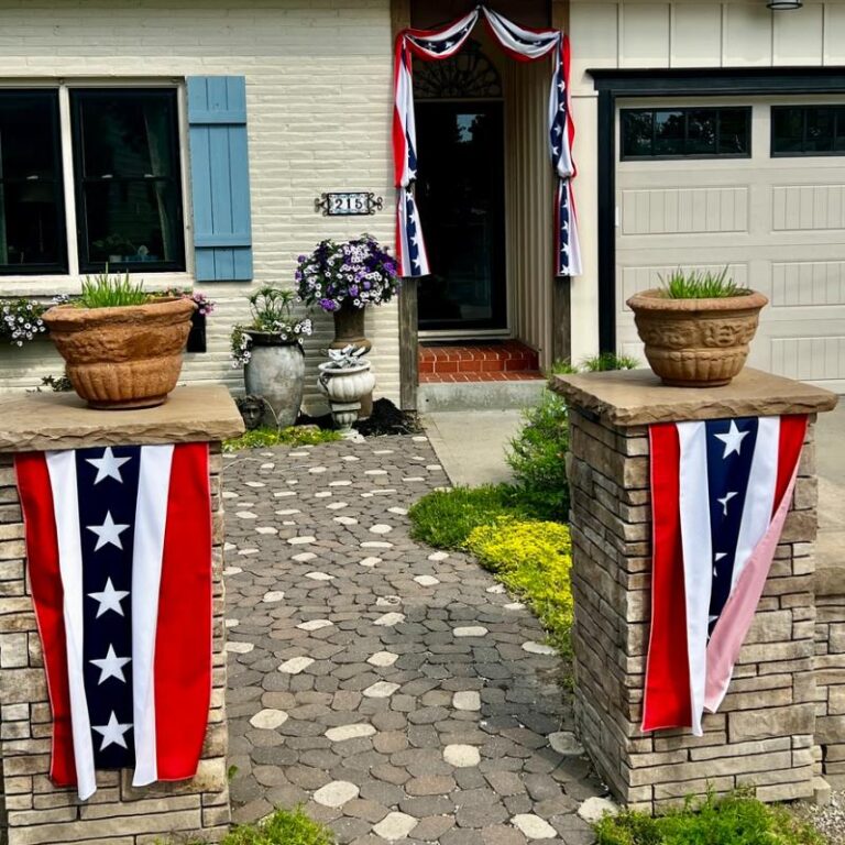 A house entrance decorated with red, white, and blue patriotic banners. Two stone pillars with star banners flank a path leading to a door adorned with similar bunting. Potted plants and flowers decorate the area.