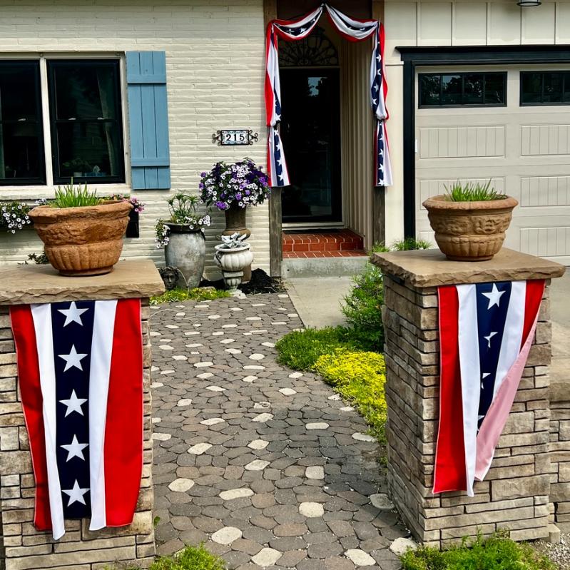 A house entrance decorated with red, white, and blue patriotic banners. Two stone pillars with star banners flank a path leading to a door adorned with similar bunting. Potted plants and flowers decorate the area.