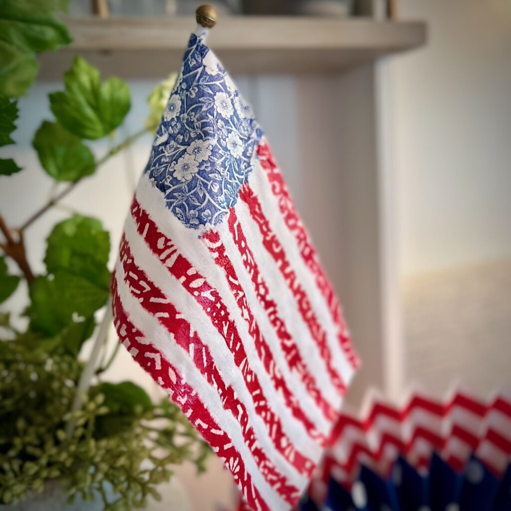 A close-up of a small decorative American flag with a floral blue field and hand-drawn red stripes, displayed indoors near green plants.