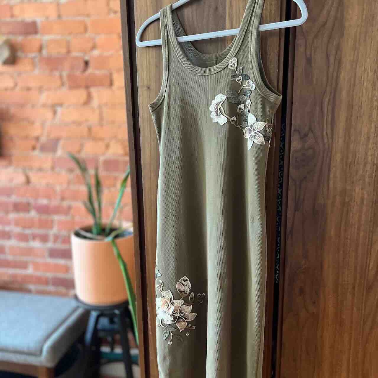 A sleeveless olive-green dress with white floral embroidery hangs on a white hanger, displayed against a wooden wardrobe. A potted plant and brick wall are visible in the background.