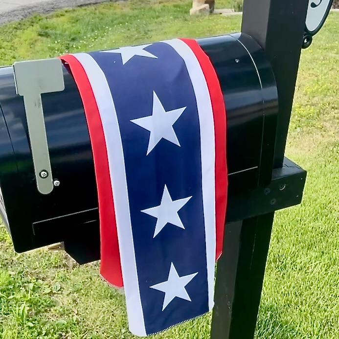 A black mailbox with a red, white, and blue ribbon featuring white stars draped over it, standing on a grassy lawn near a road.