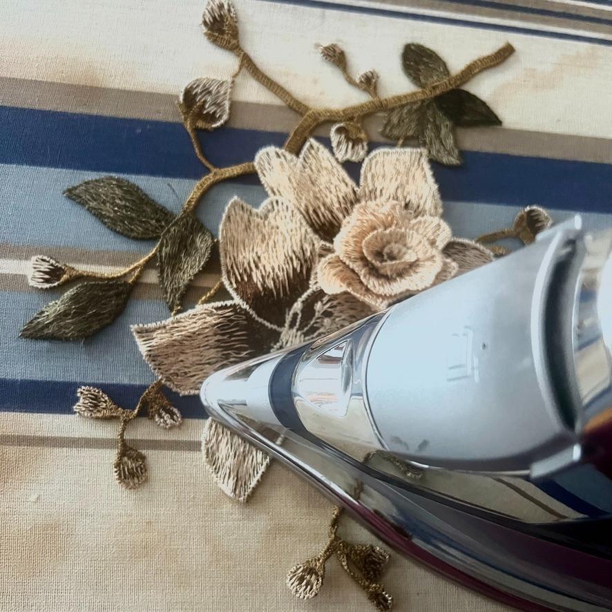 A close-up of an iron pressing a floral embroidered patch onto a striped fabric surface, with detailed brown and green flower and leaf designs visible.