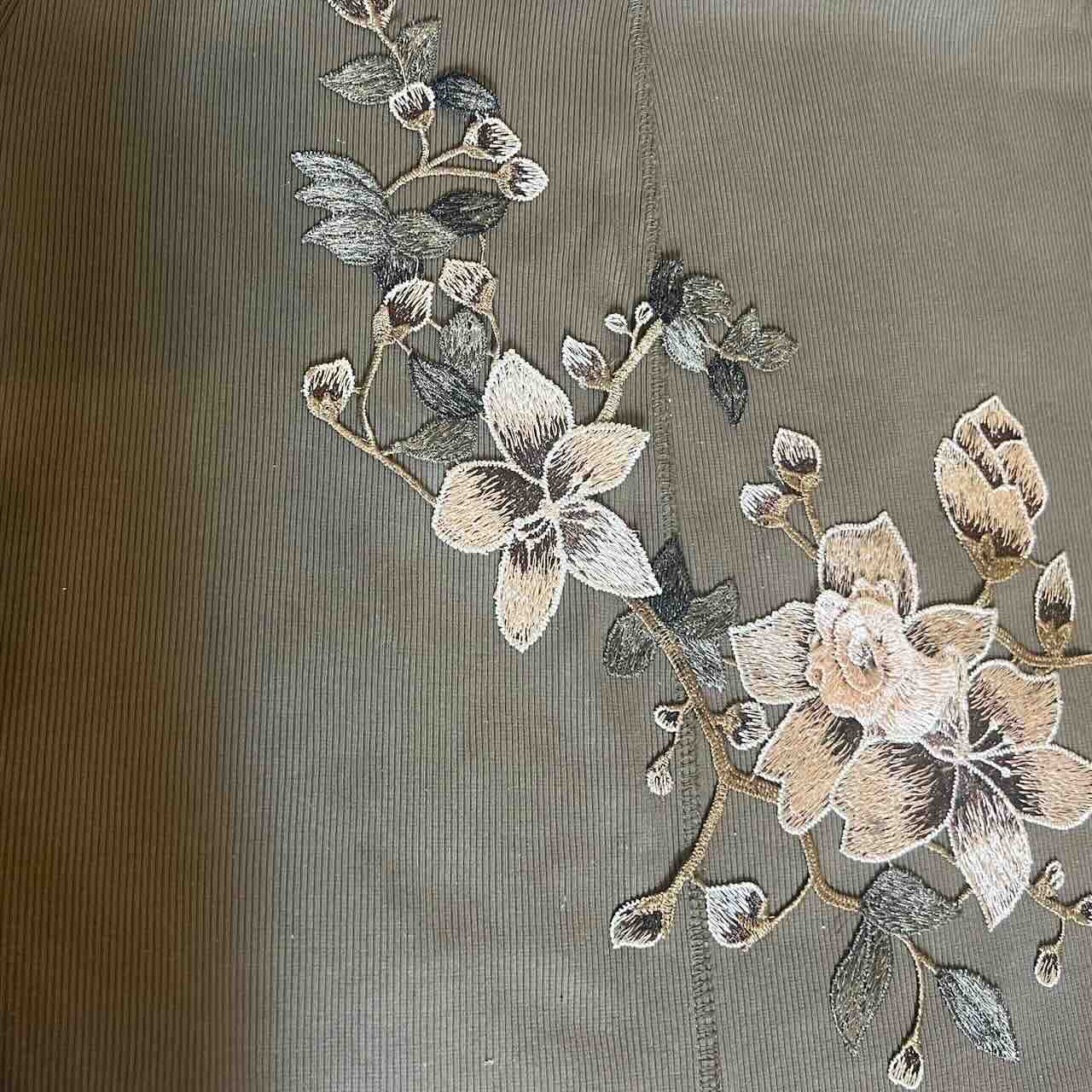 Close-up of floral embroidery featuring beige, cream, and gray flowers with green leaves on a textured, light brown fabric background.