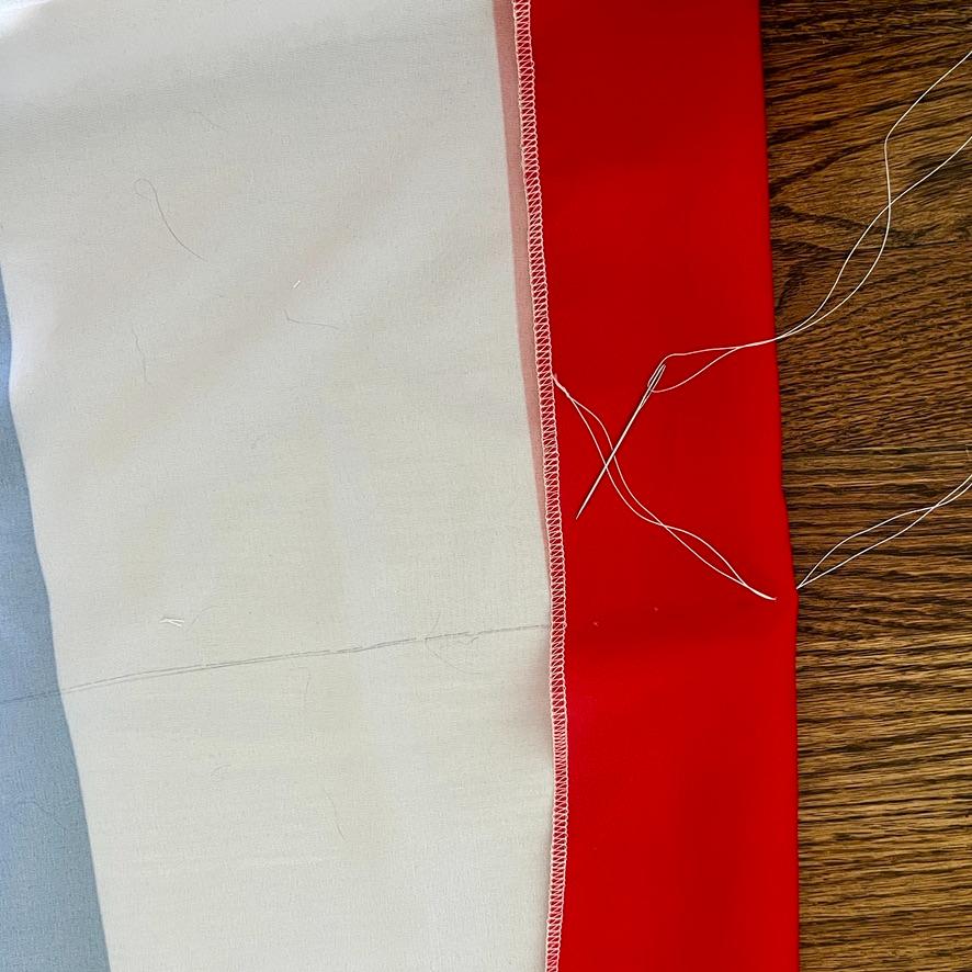 A close-up of fabric pieces—light blue, white, and red—stitched together on a wooden surface with white thread and a loose needle visible on the red section.