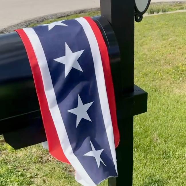 A mailbox with a red, white, and blue banner featuring white stars draped over it, standing on a green lawn near a street curb.