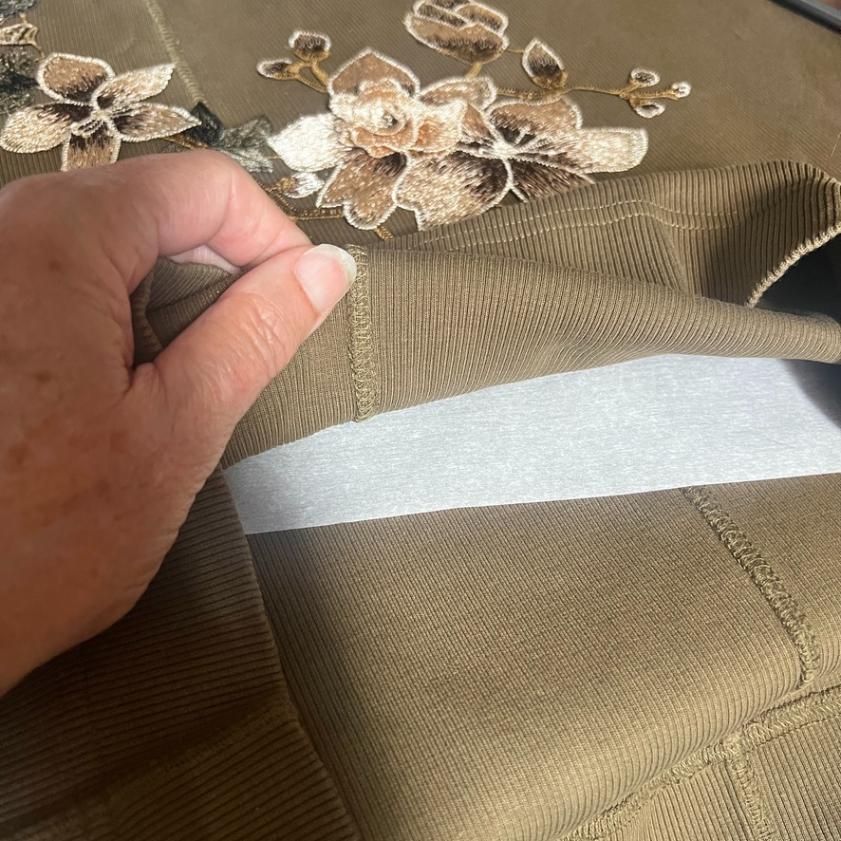 A hand lifts the hem of an olive-green garment with embroidered beige flowers, revealing a layer of white paper inside the fabric.