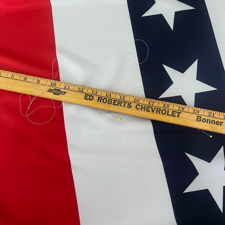 A wooden ruler with "ED ROBERTS CHEVROLET Bonner" printed on it lies diagonally on a red, white, and blue fabric with white stars. A string with yellow pins is draped over the ruler and fabric.