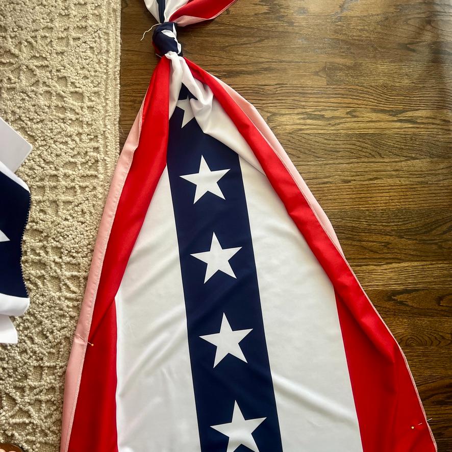 A section of a patriotic fabric with a blue vertical stripe featuring white stars, bordered by red and white stripes, lies on a wooden floor next to a textured beige carpet.