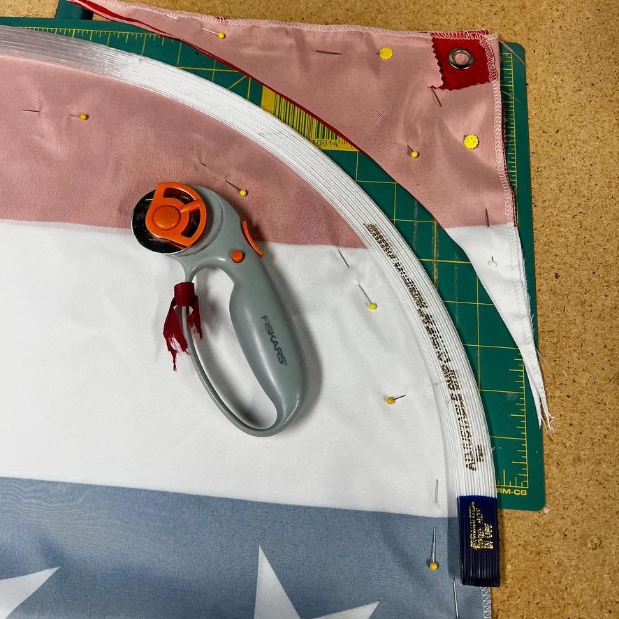 A rotary cutter rests on fabric in the colors of the American flag, placed on a green cutting mat. The fabric is pinned and partially trimmed, with a white zipper and sewing pins visible.