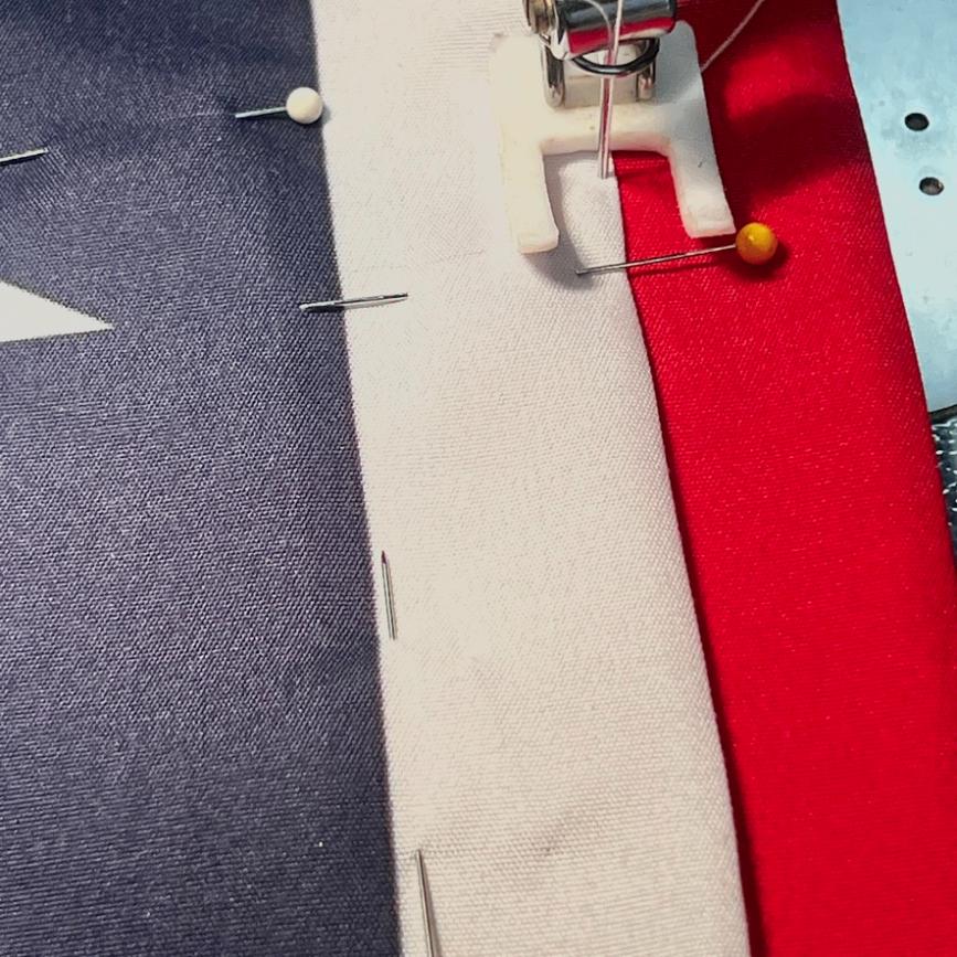 A close-up of a sewing machine stitching along the seam of a fabric that resembles part of the American flag, with pins holding the material in place.