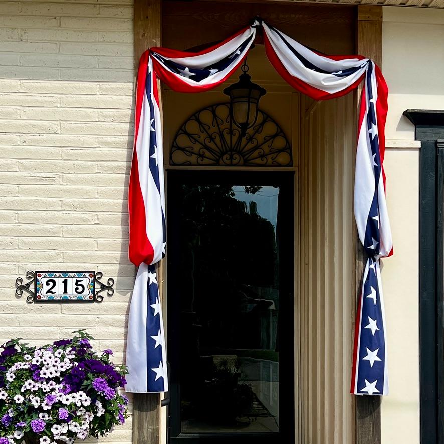 A doorway decorated with red, white, and blue bunting featuring white stars, next to a house number sign reading "215" and a pot of purple and white flowers.