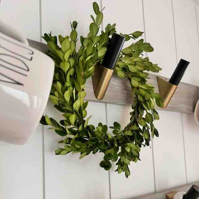 A small green wreath hangs on a wooden board with two gold hooks. A ceramic mug with black lettering is partially visible on the left side, against a white tiled wall.