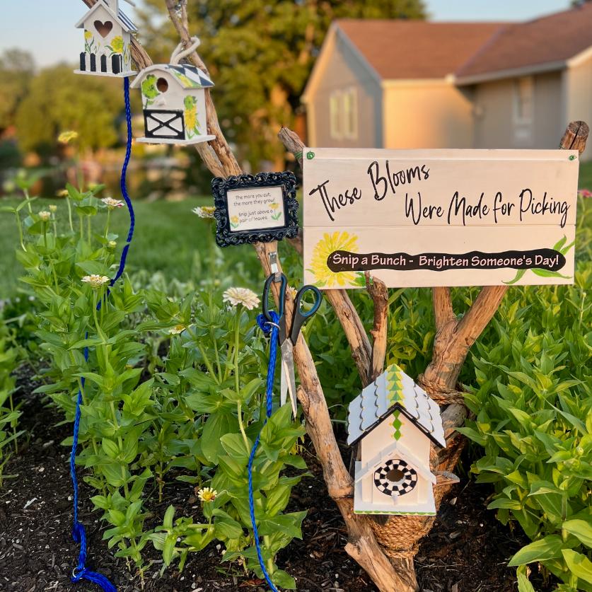 A charming neighborhood cutting garden features birdhouses perched on branches and a sign that reads, "These Blooms Were Made for Picking. Snip a Bunch - Brighten Someoneโs Day!" Vibrant flowers and quaint houses form the picturesque background.