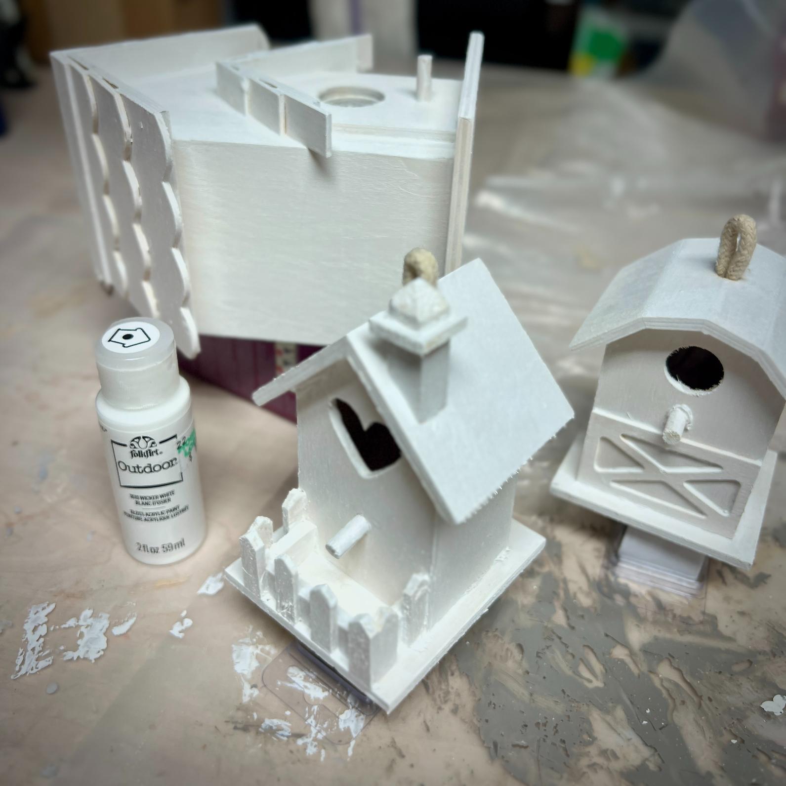 Three unpainted wooden birdhouses and a bottle of white paint are on a table covered with paint splatters and a plastic sheet, ready for a painting project inspired by the charm of a neighborhood cutting garden.