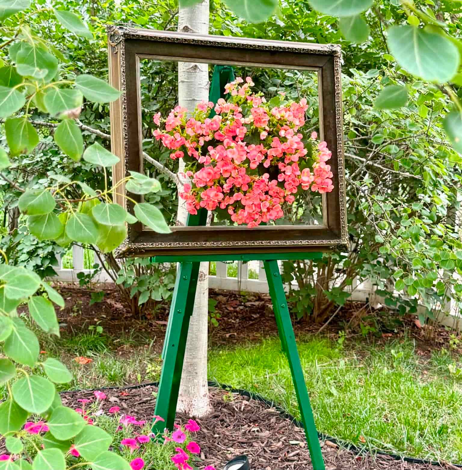 A wooden picture frame on a green easel stands outdoors in a neighborhood cutting garden, framing a cluster of pink flowers and leaves, creating the illusion of a living floral painting amid the vibrant greenery.