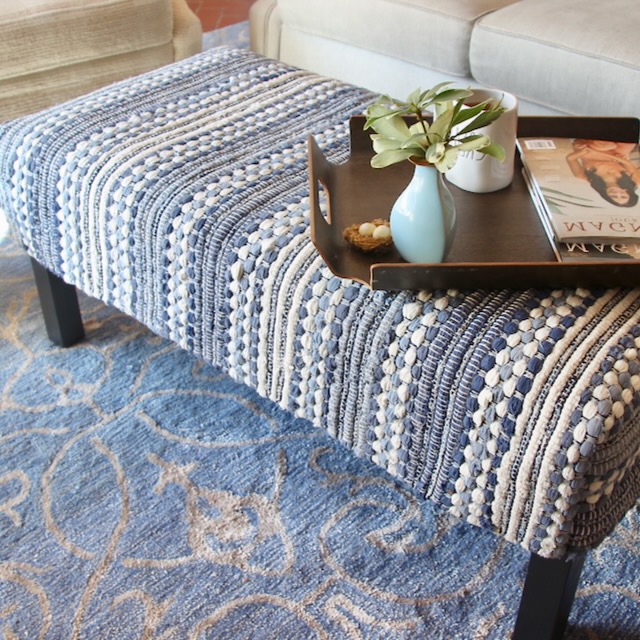 Rectangular woven blue and white ottoman on a blue patterned rug, topped with a tray holding a small vase with greenery, a mug, a cookie, and a magazine—perfect for relaxing after you Make a Folding Luggage Rack for your guests. Sofa visible in the background.