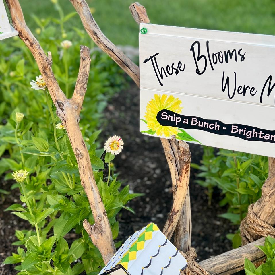 Wooden sign with yellow flower illustrations reads, "These Blooms Were Meant to Be Snipped. Snip a Bunch - Brighten Your Day!" Green plants and small flowers grow around the sign, inviting all to enjoy this charming neighborhood cutting garden.