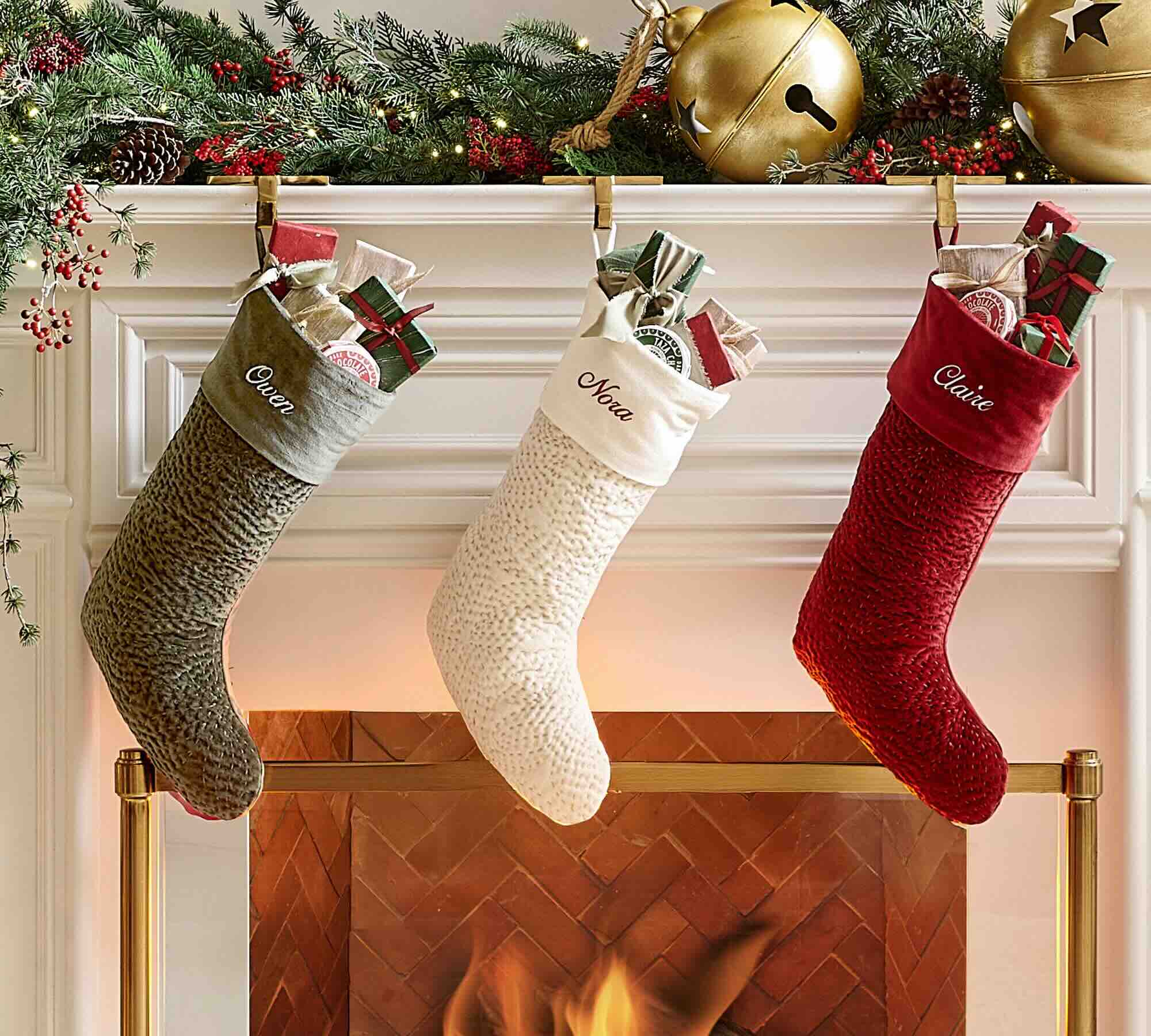 Three Christmas stockings labeled Owen, Nora, and Claire hang above a fireplace, filled with gifts. Greenery and gold bells decorate the white mantel, and a fire glows warmly below.