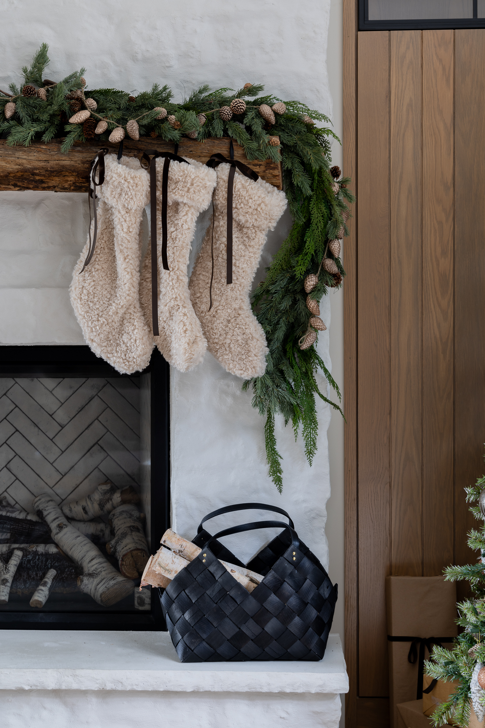 Three fluffy stockings hang from a mantel decorated with pine garland and pinecones. Below, a black woven basket holds firewood beside a fireplace, with part of a Christmas tree in the corner.