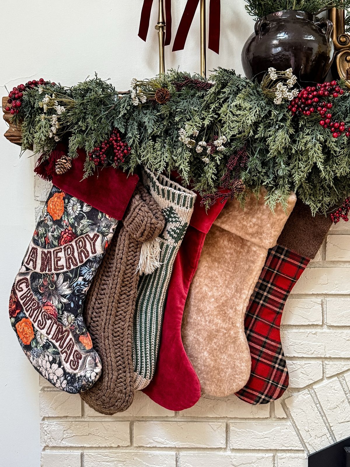 Five Christmas stockings in different patterns hang from a decorated mantel with greenery and red berries above a white brick fireplace.