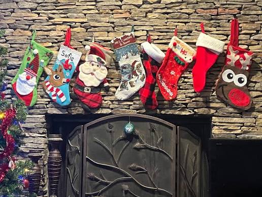 Seven colorful Christmas stockings with festive designs hang on a stone fireplace. There is a decorated Christmas tree on the left and a black iron fireplace screen below the stockings.