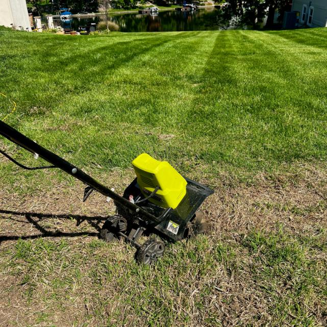 A yellow and black tiller sits on a patch of dry grass, with a green, freshly mowed lawn and a lake with boats visible in the background—perfect scenery for maintaining your neighborhood cutting garden.