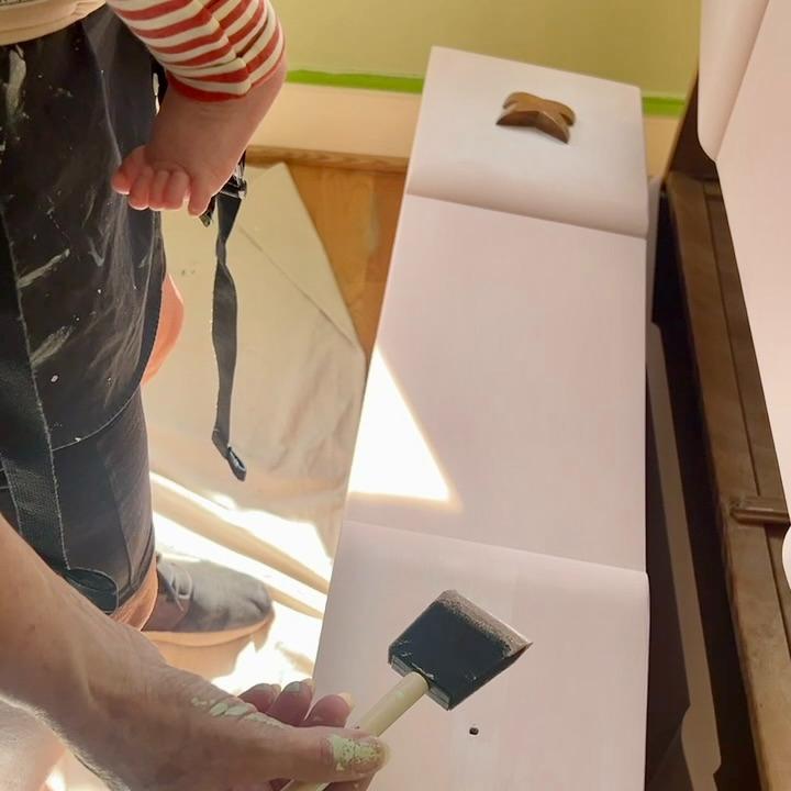 An adult holding a baby paints a white surface with a foam brush; the adult’s shirt and shorts have paint stains, and the baby wears striped sleeves.