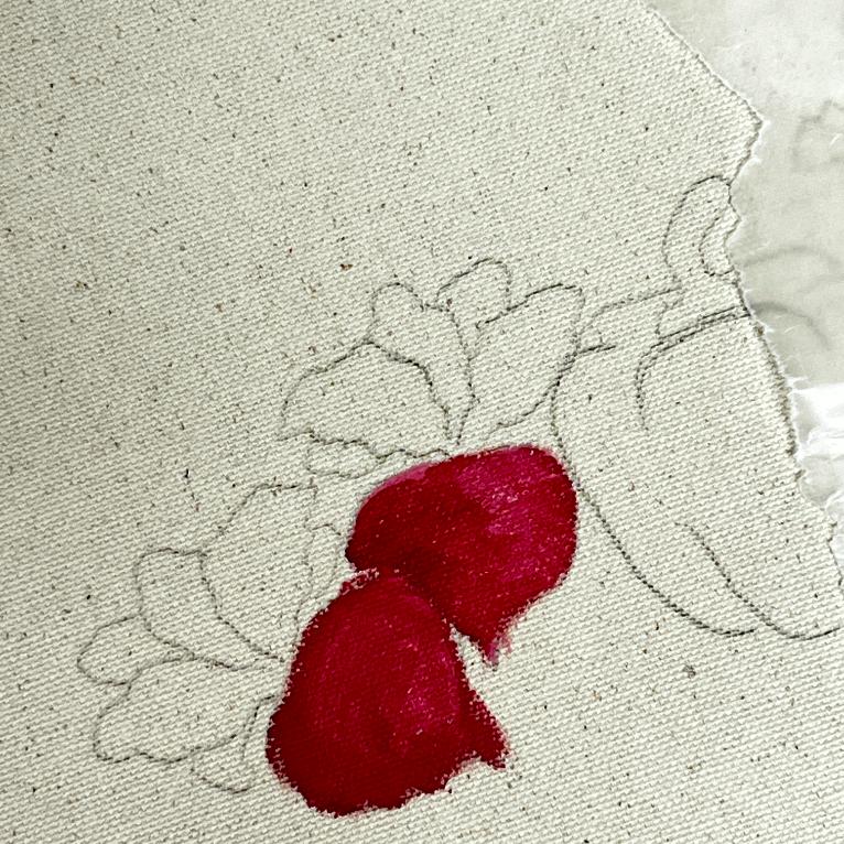 A piece of canvas with a floral outline, partially colored in with two bright red flower petals. The rest of the design remains uncolored, and the fabric edges are slightly frayed on the right side.
