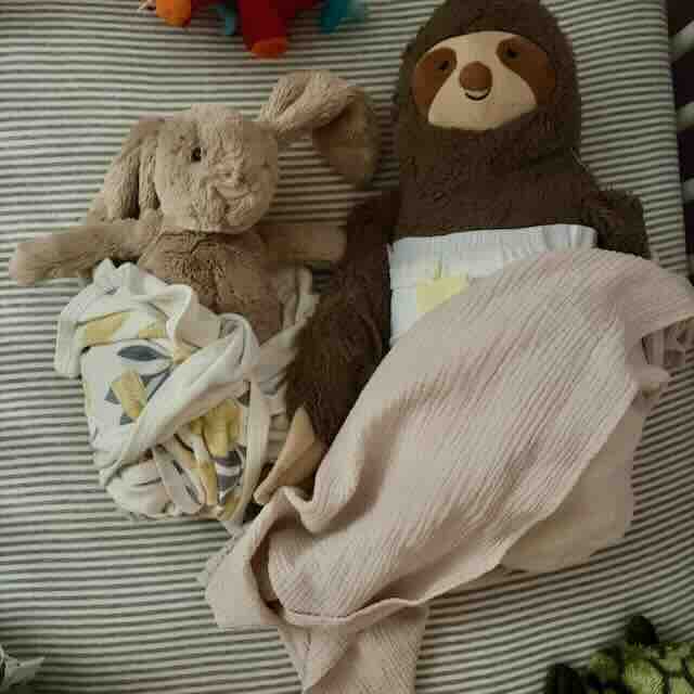 Two stuffed animals, a bunny and a sloth, are wrapped in diapers and blankets, lying side by side on a striped surface as if tucked in to sleep.