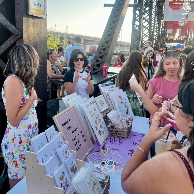 A group of women browse handmade cards and jewelry at an outdoor market booth, chatting with the vendor under sunny skies near a metal bridge structure.