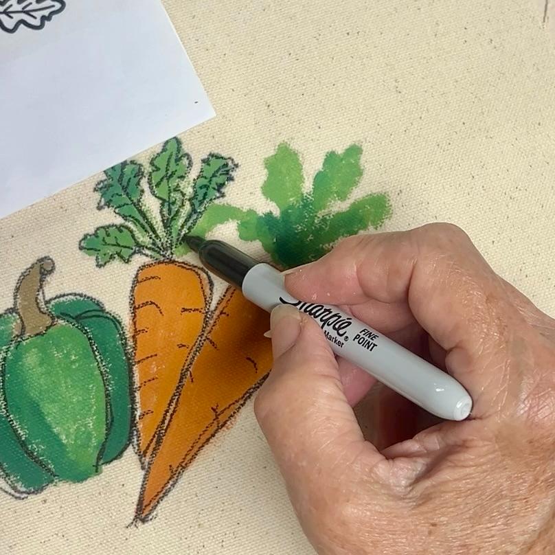 A hand uses a black Sharpie marker to outline colorful drawings of vegetables, including carrots and a green bell pepper, on a canvas. A piece of paper with part of the drawing is visible in the corner.