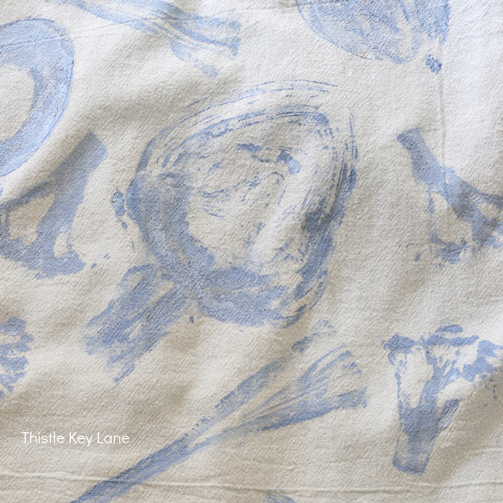 A white cloth with abstract blue prints resembling vegetables, such as artichokes and broccoli. The text “Thistle Key Lane” appears in the lower left corner.