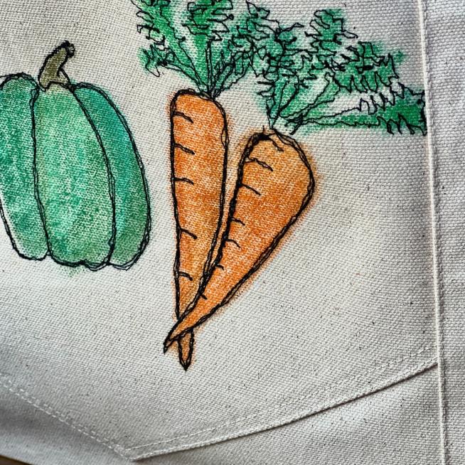 Hand-drawn illustration of a green bell pepper and two orange carrots with green tops on beige fabric, possibly part of a tote bag or apron.