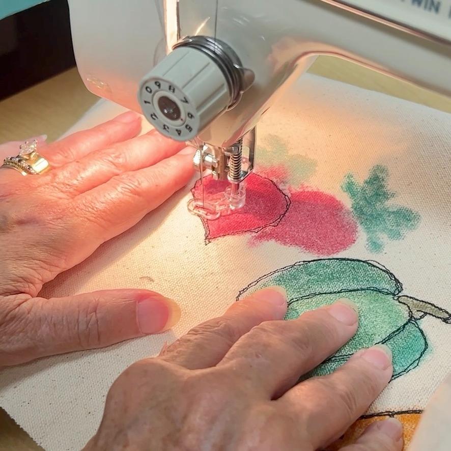 A person guides fabric with colorful vegetable illustrations under a sewing machine needle, stitching details onto the designs.