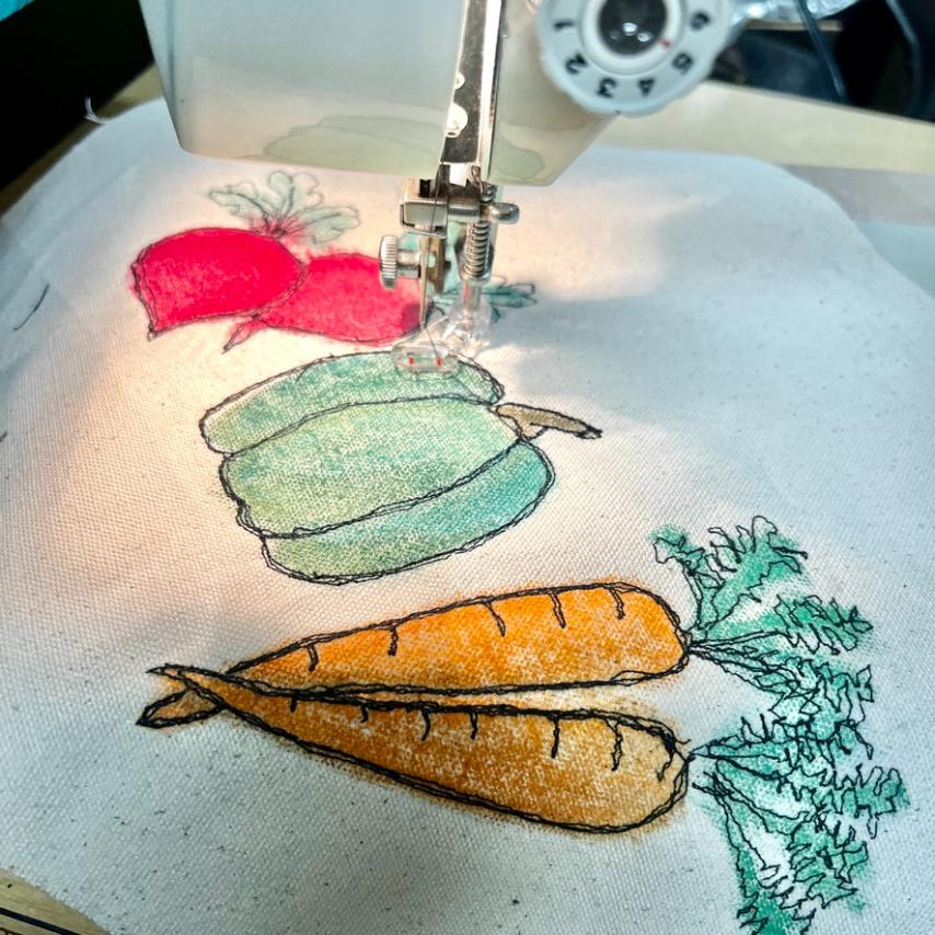 A sewing machine stitches colorful vegetable designs—beet, squash, and two carrots—onto white fabric, with visible thread outlining and shading each vegetable.